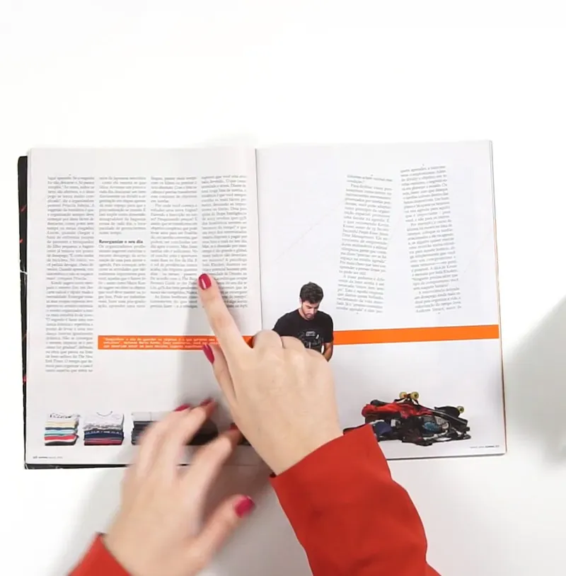 Mãos folheando uma revista aberta, com textos, fotos e uma faixa laranja