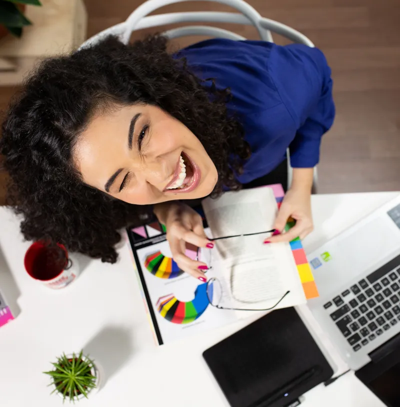 Designer sorrindo, vista de cima, numa mesa com leques de cor, caderno e notebook
